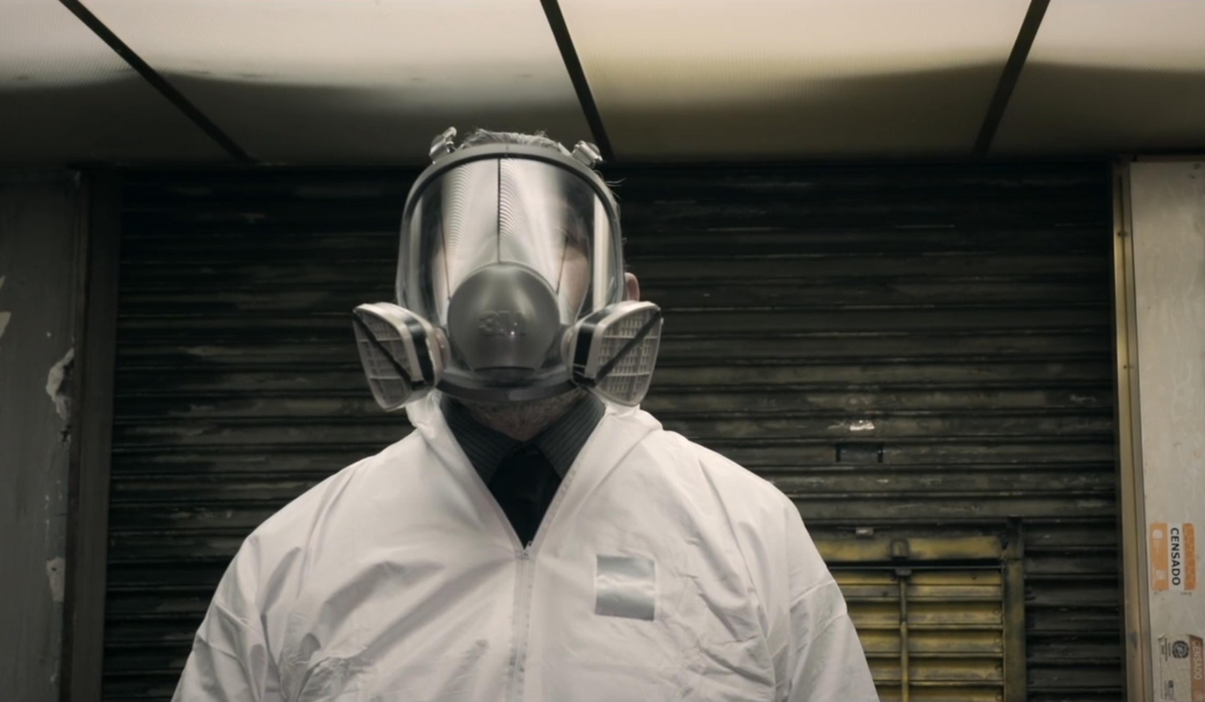 Person wearing a white hazmat suit and a full-face respirator, standing in a dimly lit industrial setting.
