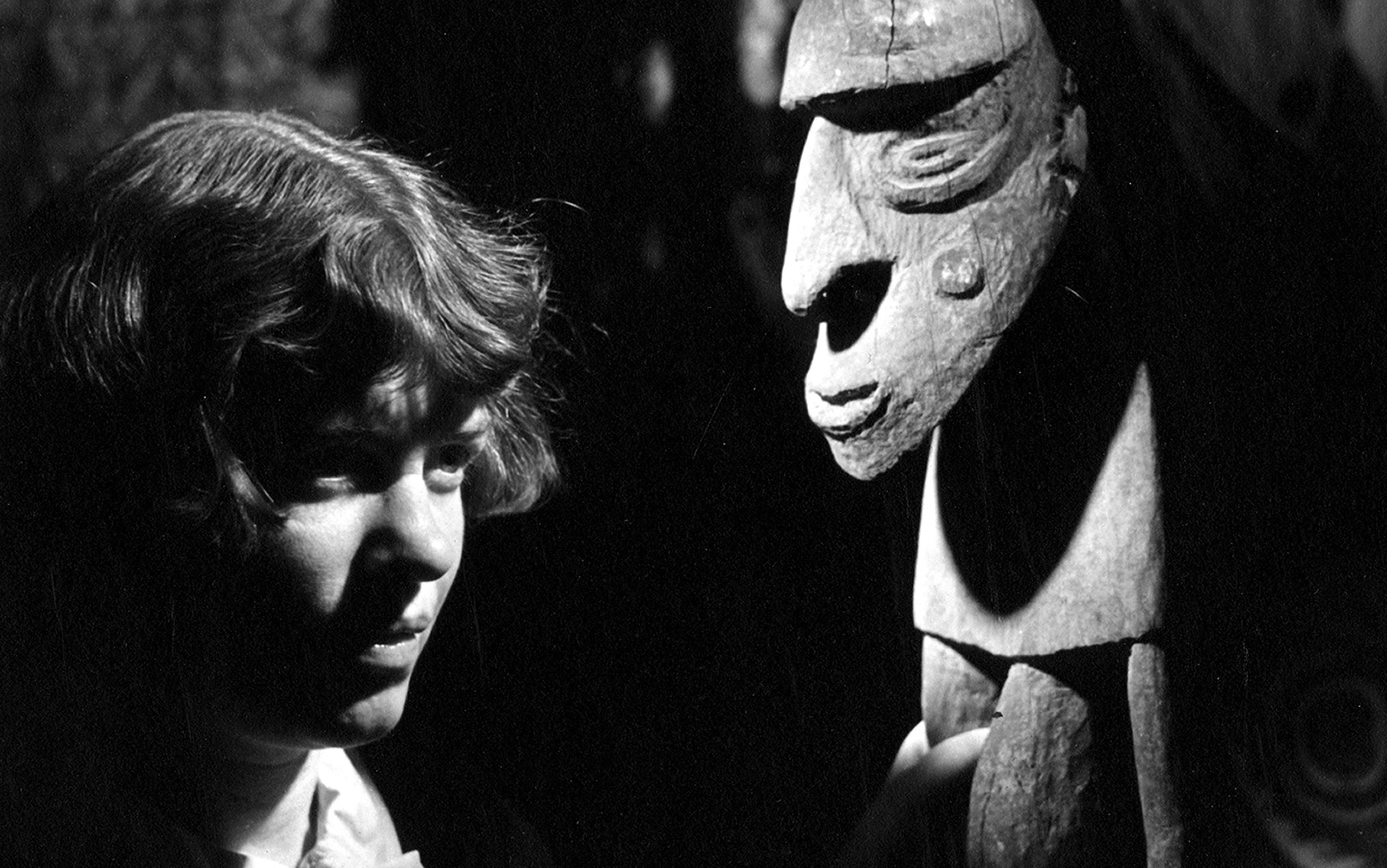 Vintage photo of a woman with short hair who is Margaret Mead looking at a carved wooden tribal figure against a dark background.