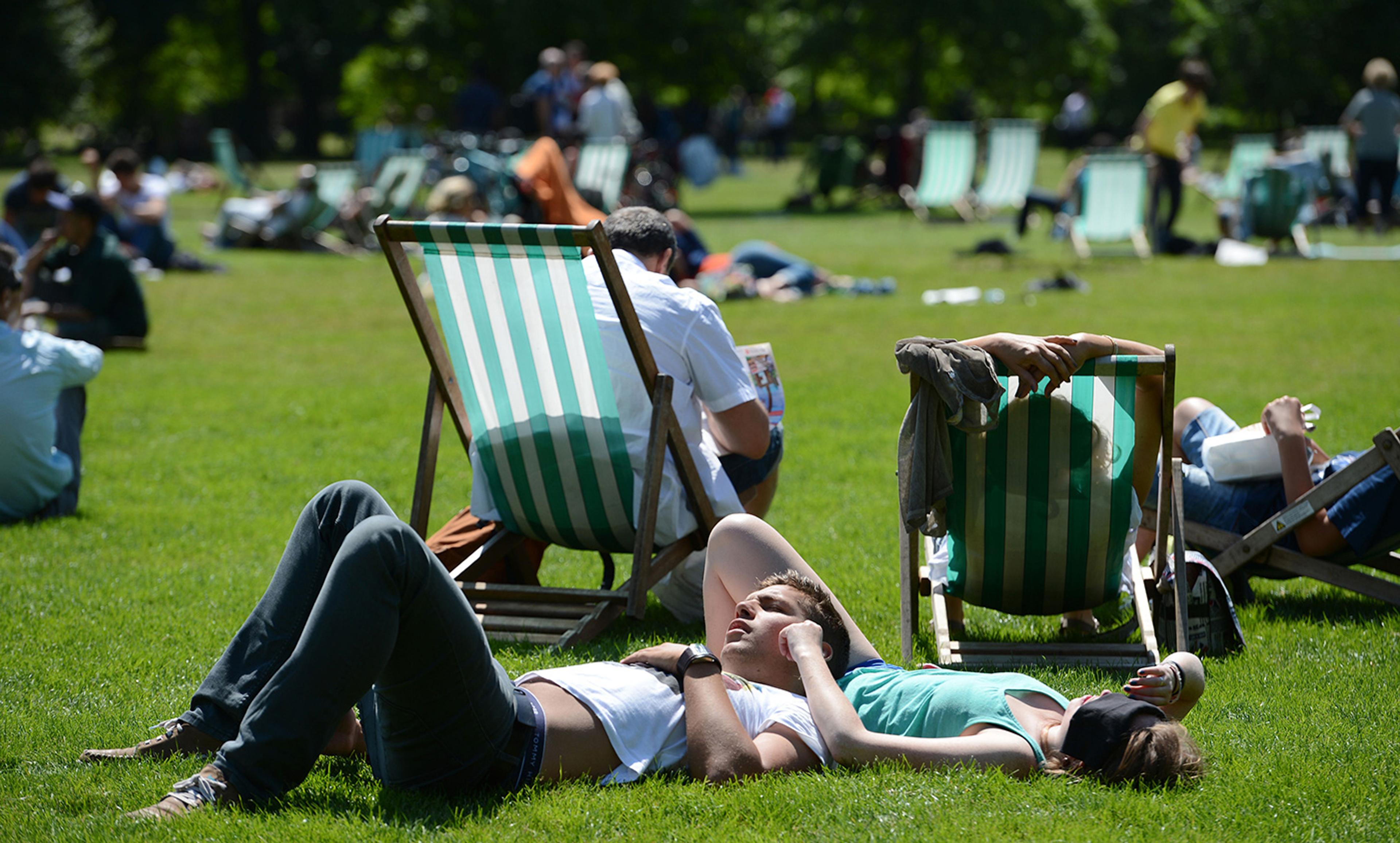 People relaxing on green-striped deckchairs and grass in a sunny park, with trees in the background and more people seated.